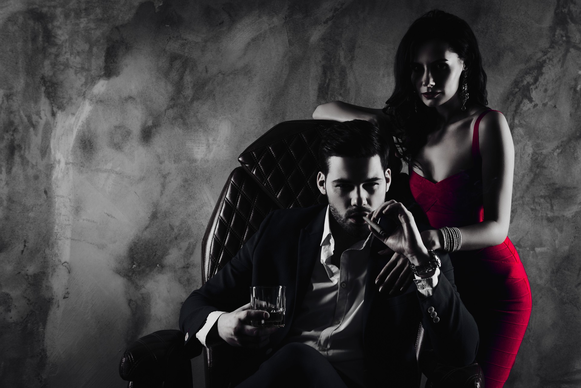 Portrait of brutal gentleman in formal wear sitting in leather chair holding glass with whiskey cigar in hands, charming pretty lady standing near looking at camera isolated on grey background